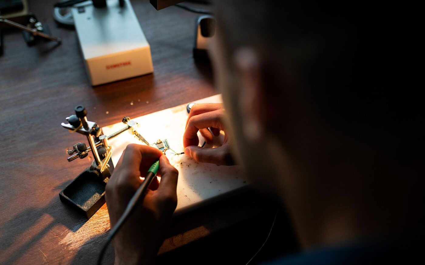 Engineer hand-soldering a small circuit board under a bench lamp.