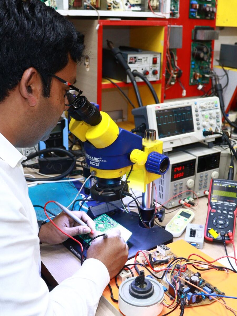 Technician probing a circuit board under a stereo microscope at a bench fitted with an oscilloscope and programmable power supply.