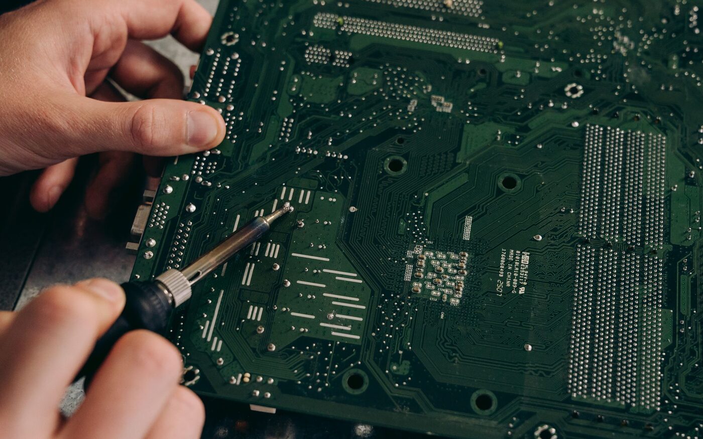 Hands using a soldering iron to work on the underside of an existing circuit board.
